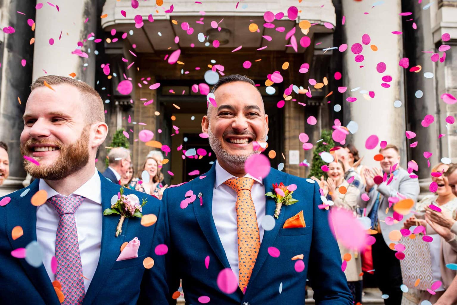 two smiling grooms having pink and white wedding confetti thrown at them after their wedding