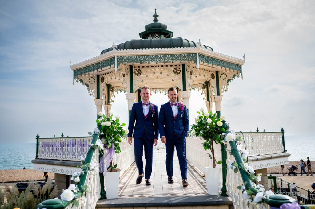 two grooms smiling and walking down Brighton Bandstand