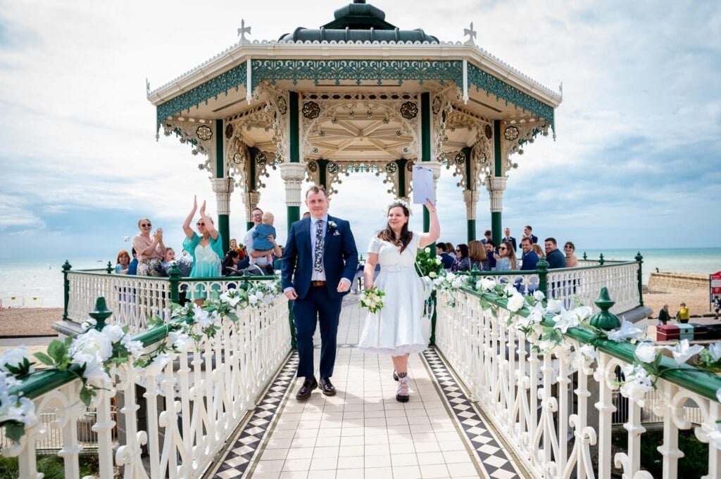 couple waving their marriage certificate, having just got married at the Brighton Bandstand their Brighton Beach Party Wedding to follow